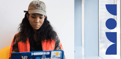 Une femme concentrée travaillant sur un ordinateur portable recouvert de stickers liés à la tech et à la diversité, assise dans un bureau moderne.