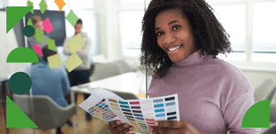 Une femme souriante tient une palette de couleurs dans un bureau lumineux, tandis que des collègues discutent devant un tableau recouvert de post-it colorés.